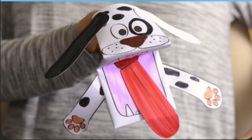 Mão segurando e manipulando um fantoche de papel dobrado em formato de cachorro dálmata, com boca aberta e língua de papel saindo